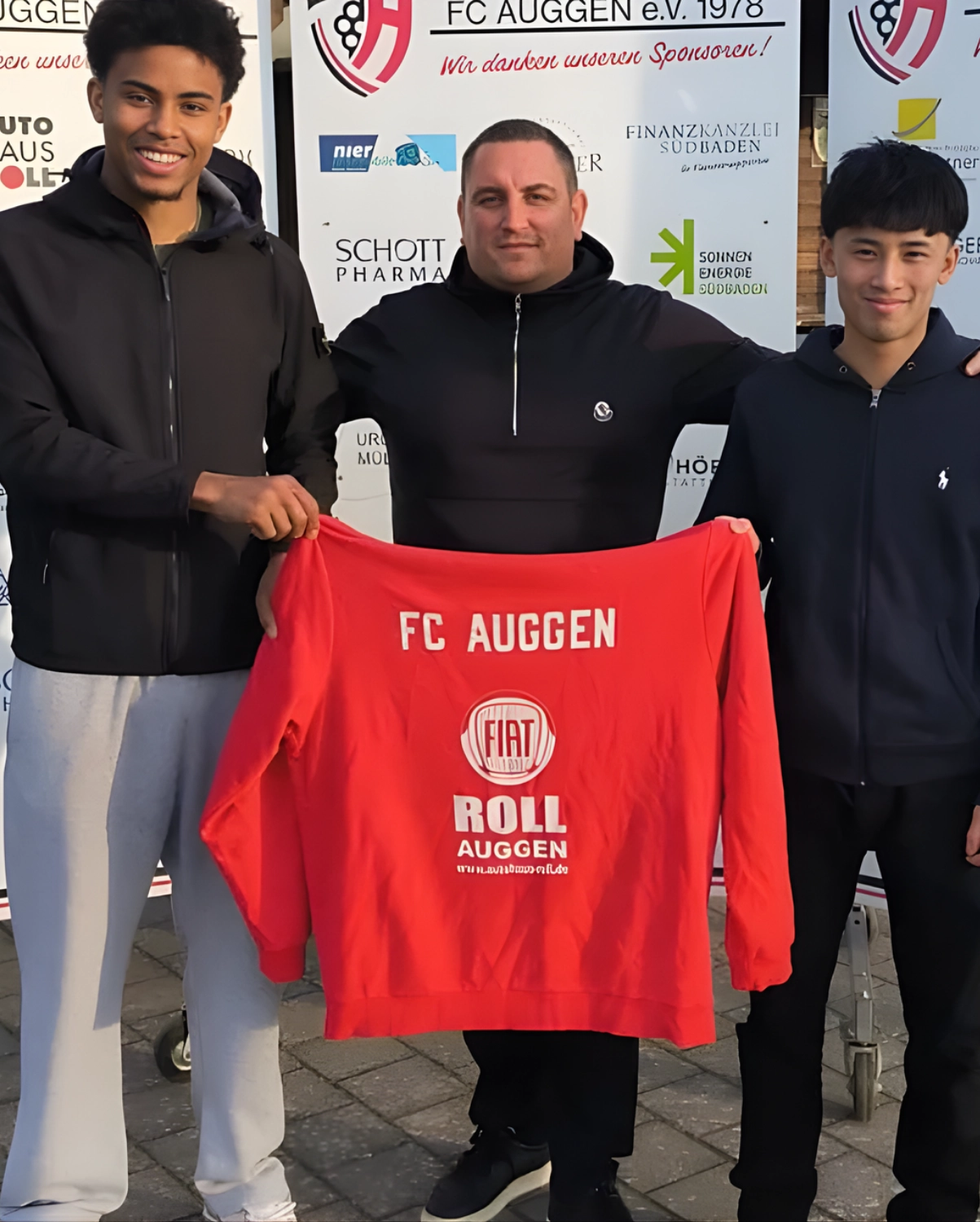 FC Auggen e.V. – Fußballtrainer – Dieses Bild zeigt drei Personen, die vor einer Sponsorentafel ein rotes Sweatshirt mit den Aufschriften „FC AUGGEN“ und „FIAT ROLL AUGGEN“ hochhalten. Leider konnte ich keine weiteren Details angeben, da die Suche keine hilfreichen Informationen lieferte.